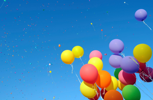 A large collection of colorful balls or balloons flys through the streets of a city. They fly towards the sky and pass by large buildings. It is a surreal sight.