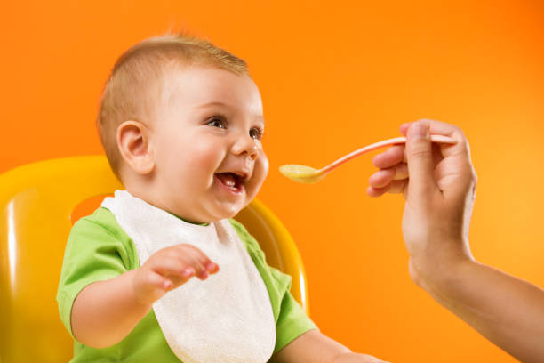 Feeding cheerful baby on orange background Mother's hand feeding a laughing baby in front of a vivid orange background. baby spoon stock pictures, royalty-free photos & images