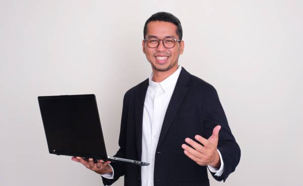 istock Asian businessman smiling and showing inviting hand gesture while holding a laptop 1871045028