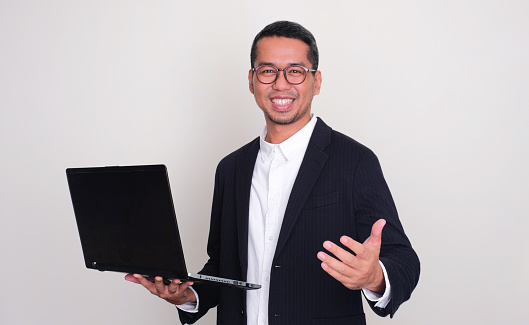 Asian businessman smiling and showing inviting hand gesture while holding a laptop