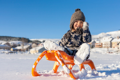 Cheerful boy in winter clothes sitting on sled in snowy field, celebrating successful downhill sledding. Snow is on his hands and legs, winter scenery with mountain village and crisp blue sky is in the background.