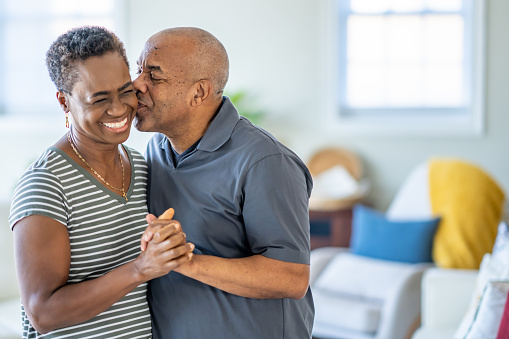 A senior couple of African ethnicity, are seen dancing together in the comfort of tehir own home.  They are both dressed casually and are smiling as they enjoy each others company and the time to bond closer together.