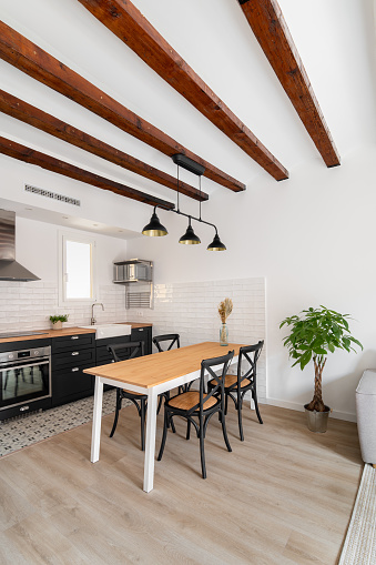 Dining table with wooden surface and chairs set in kitchen room. Ceiling decorated with wooden beams. Place to eat and rest with cooking appliances in renovated house