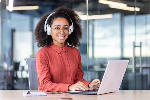 Portrait of young beautiful successful woman at workplace inside office, businesswoman in headphones smiling looking at camera, satisfied with achievement results programmer with laptop.