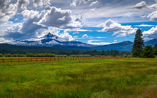 Mount Washington in Oregon with a vibrant green field and wooden fence under a dramatic cloudy sky. Mount Washington is a deeply eroded volcano in the Cascade Range of Oregon, USA.