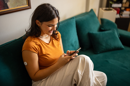Young diabetic patient teenage woman using mobile phone while sitting on sofa at home, she has a glucose monitor on her arm.
