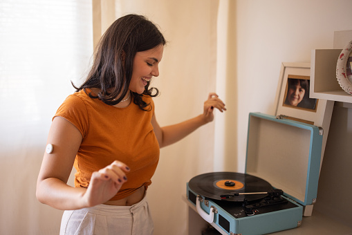 Young diabetic patient teenage woman listening music on turntable and dancing at home, she has a glucose monitor on her arm.