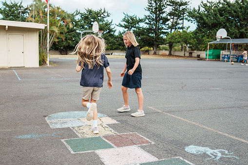 A happy and active schoolgirl of Caucasian ethnicity plays hopscotch with her friend during an outdoor recess on a cloudy Fall day.