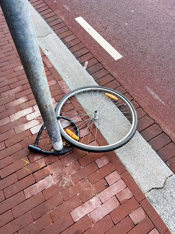 A lone bicycle wheel, once belonging to a forgotten ride, stands locked to a sturdy street pole. Its spokes glisten in the daylight while the empty frame tells a tale of abandonment. Dutch culture background, copy space.