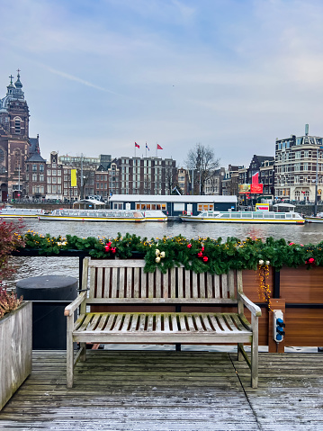 Beautiful Christmas decorated bench and view of the Amstel river and surrounding buildings from Amsterdam Centraal on a cold grey winter day in December. Boats leaving for tourist tours. Dutch culture and destination background, copy space.