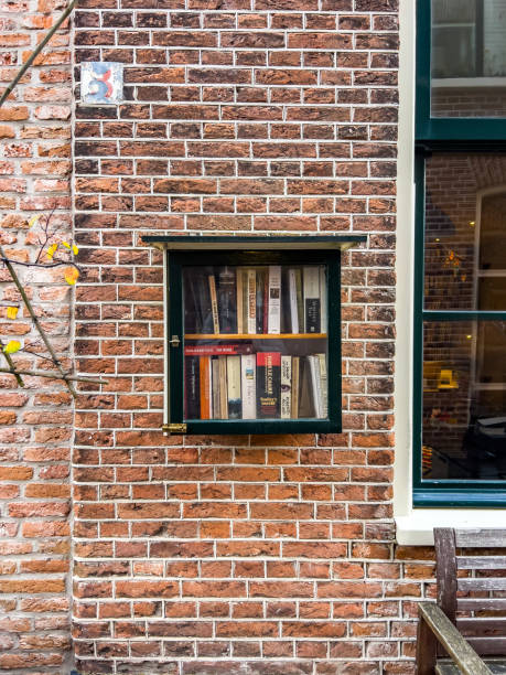 A square, locked box with a window frame, filled with books, is hanging outside on the brick wall of a house. Cities; Haarlem street scene.