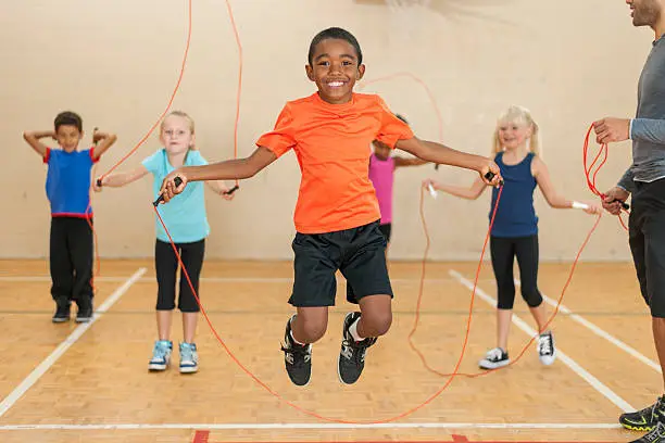 Smiling boy is skipping rope.  The boy in the foreground is shown while jumping in the air, and there are three other children with ropes in the background.  There is an adult on the right side of the photo.