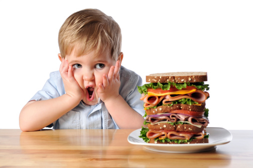 Cute image of a little boy with a very large sandwich. It is layered with deli meats, cheeses, purple onion, tomato and lettuce on wheat bread. The boy has a very shocked look on his face. How will he be able to eat all of it?
