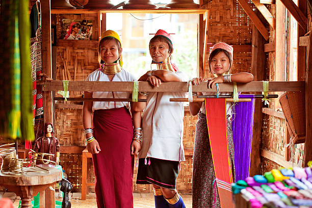 Attractive Kayan, or Padaung, 'long neck' young tribal women with heavy neck rings  working at textile weaving looms in open hut, Inle, Myanmar, Southeast Asia. Myanmar, formerly Burma, is a predominantly Buddhist culture with a plethora of shrines, temples and places of worship distributed throughout the country, which is a colorful amalgam of the original Tibeto-Burman and Pagan kingdoms that now show little sign of British and Japanese rule but now notable for its precious stones, tourism and dubious human rights record.