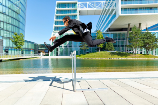 young hurdler in business suit running over hurdle in modern office complex outdoors at sunshine