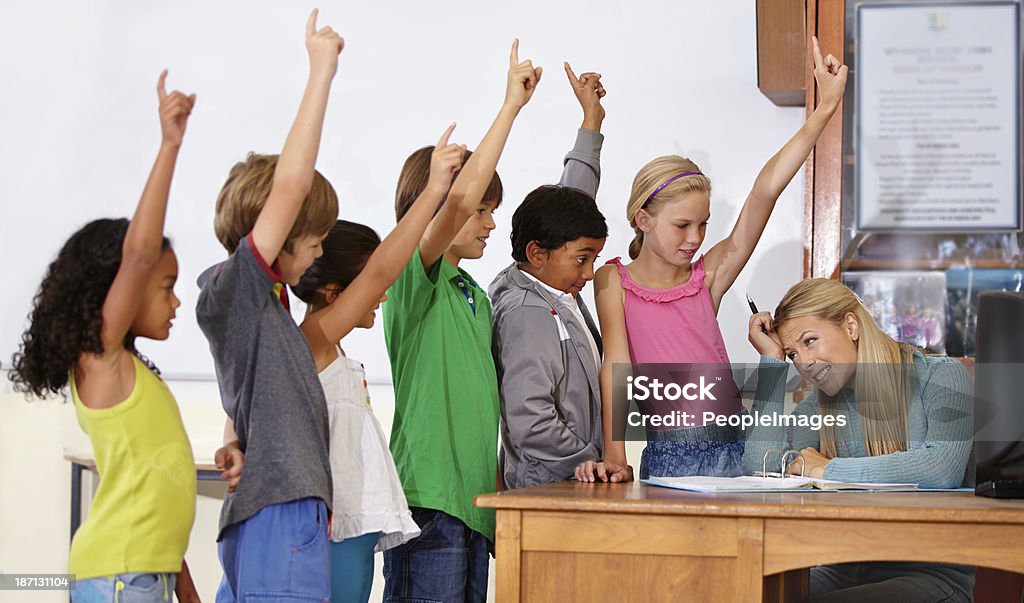 We know the answer! A teacher sitting at her desk talking to her students who all have their hands up Adolescence Stock Photo