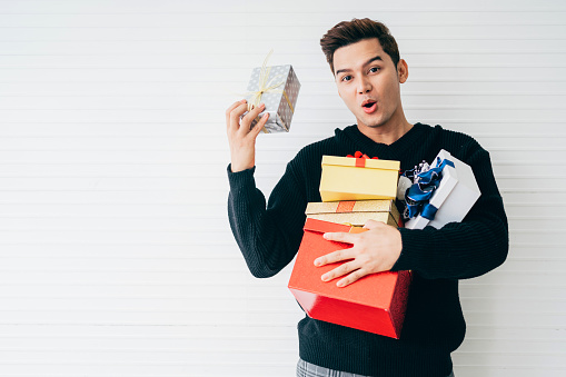 Portrait of an excited and surprised young Asian man wearing Christmas sweater holding many gifts that stacking in his hands in the living room with white background. Image with copy space.