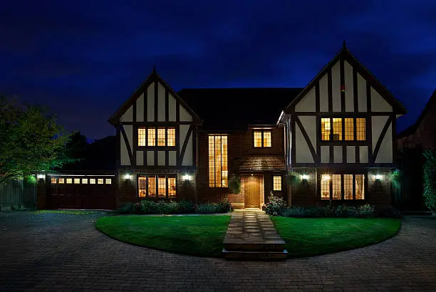 an evening view of a luxury mock-Tudor home taken a little while after sunset in order to expose the deep blue sky. All of the light of the house have been turned on. Bright white external lights at the front of the house and pillar lights (out of view) around the driveway have also lit up the lawn. This is NOT an HDR image. 