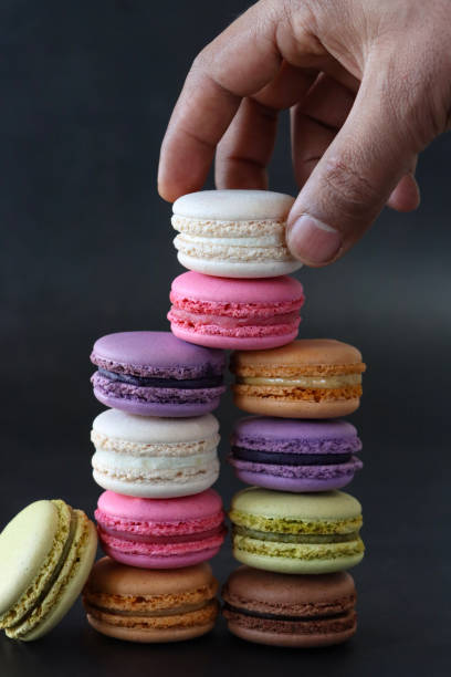 istock Image of unrecognisable person building stacks of multi coloured macarons filled with flavoured butter cream, pistachio macaron leaning against blueberry, strawberry, coffee, chocolate flavoured meringues, black background, focus on foreground 1871519657