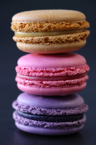 Stock photo showing close-up view of a stack of multi coloured macarons on a black background. Pink strawberry, brown chocolate, and purple blueberry flavoured meringues.