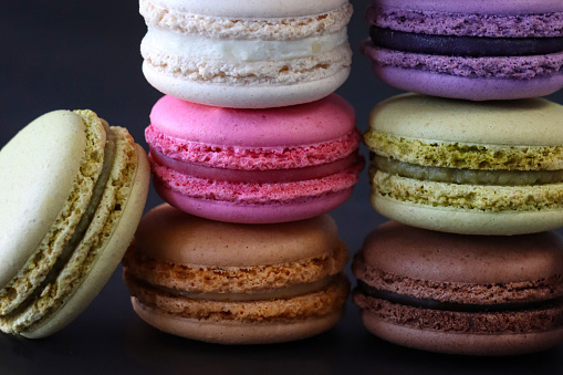 Stock photo showing close-up view of stacks of multi coloured macarons on a black background. Pink strawberry, brown chocolate and coffee, green pistachio, white vanilla and purple blueberry flavoured meringues.