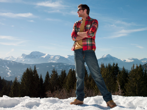 A man stands on the snowy mountain looking at the view with the landscape of mountains behind him.