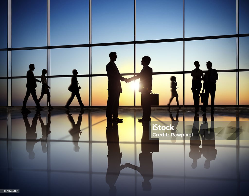 Silhouette people in office building shaking hands Business people walk and meet in a large corridor of an office building.  The scene is against a sunny sky behind a large office window.  The people appear as silhouettes and are reflected off of the surface of the floor. In Silhouette Stock Photo