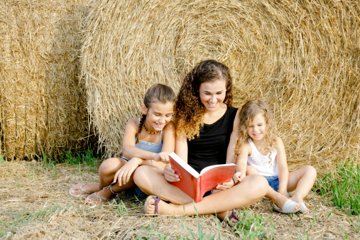 Three sisters, from toddler to teen, reading a book in the country.