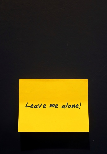 A yellow note with text written LEAVE ME ALONE ,stick on black wall background. Concept of  refrain from disturbing, interfering with someone, need private time.