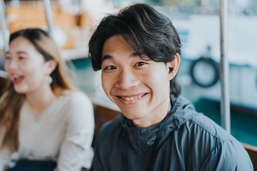 A close-up shot of a young Chinese male tour guide smiling brightly towards the camera. He is wearing an earphone. The woman beside him in white is also laughing gracefully as they embark on their Sampan boat adventure.