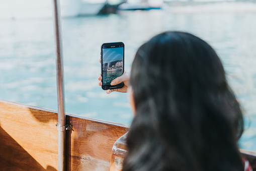 A close-up of a female tourist taking a picture of the spectacular ocean view from the Sampan boat. This shoot focuses on capturing the picture taken on the mobile phone. There are vibrant cities, blue clean ocean, and white ships.