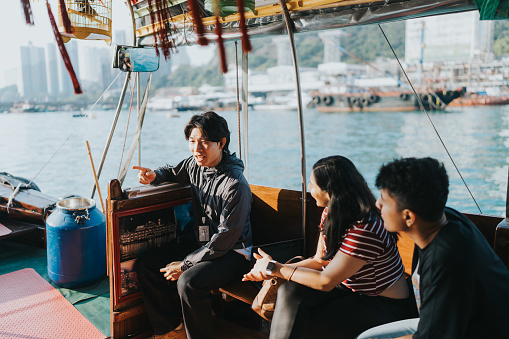 A medium shot of a young Chinese male tour guide explaining about the Sampan boat with his hand pointing at something. The Indian tourists actively listen to his explanations. They are riding the Sampan boat happily.