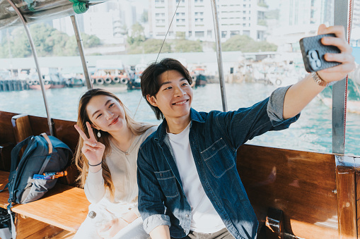 A medium shot of a young Chinese couple taking a selfie together on the Sampan boat. The boyfriend is holding a black phone to capture their moments together. The girlfriend is smiling while making a peace sign. The background is filled with various boats and ships, making the view more breathtaking.