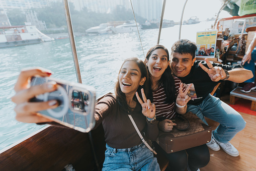 A medium shot of two Indian female tourists and an Indian male tourist taking a selfie together on the Sampan boat. All of them are smiling happily with their hands doing a peace sign. They are capturing the breathtaking moments and turn them into memories.