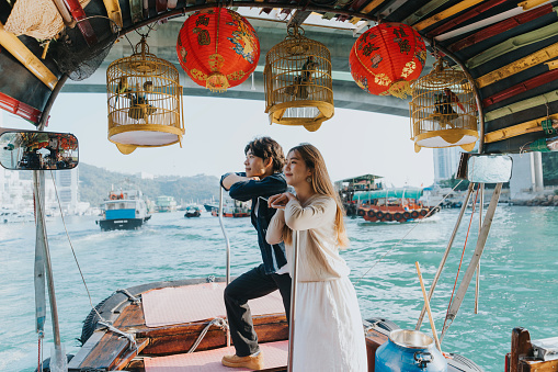 A medium shot of a young Chinese couple leaning on the railing while looking at the wonderful ocean view. Above them are some Chinese lanterns and birdcages. They are smiling gracefully gazing at the boats passing by.