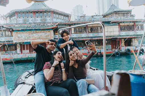 A medium shot of a group of multicultural tourists taking a selfie together in front of the Aberdeen's fishing harbor on the Sampan boat. The woman in brown sweater is holding the white mobile phone while making a thumbs up pose. The Indian tourist beside her and Chinese man behind her are also making a thumbs up pose while smiling. The Chinse tourist in white is making a cute face and a peace sign. The Indian man in black is making a peace sign in front of his forehead.