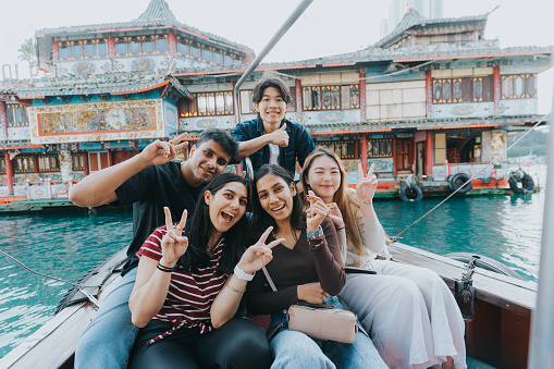 A medium shot of young multicultural tourists smiling towards the camera. The female tourists in stripes and white are making peace sign poses. The woman in the middle is making a heart-shaped pose with her left hand. The man in black is also making a peace pose, while the man in blue and white is doing a thumbs up. In the background is the Aberdeen's fishing harbor.