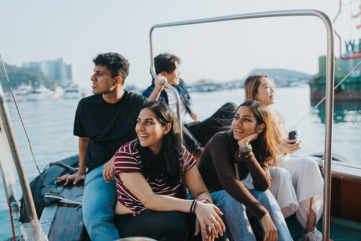 A medium shot of a group of young multicultural tourists enjoying the ocean view from the Sampan boat. The Indian tourists are looking to the right side while laughing at something. The Chinese tourists in the background are smiling while looking in the opposite direction