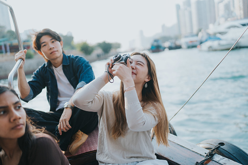 A medium shot of a young Chinese female tourist taking a photo of the sky using a camera. She smiles as she takes the picture. The male tourist behind her is sitting cross-legged with his hand on the railing