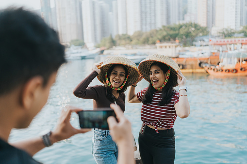 A medium shot of a male foreign tourist taking a phone photo of two international female tourists wearing traditional Hong Kong hats. In the background, sampan wooden boats and skyscrapers adorn the view