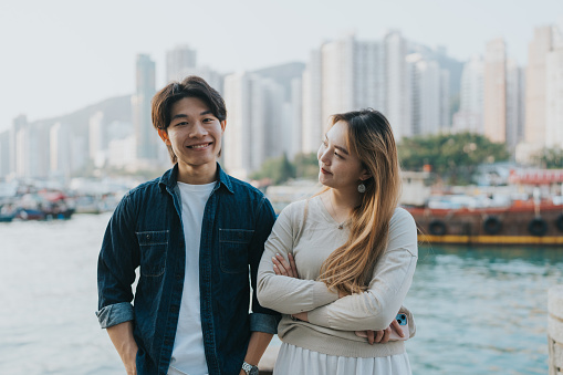 A medium shot of a smiling young Chinese couple. The girlfriend is adoringly looking at her smiling boyfriend with crossed arms. In the background, local Sampan wooden boats and skyscrapers adorn the view