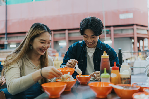 A medium shot of a smiling young Chinese couple. They are eating Hong Kong local food. Multiple small orange bowls with different delicacies and sauces are scattered around the table