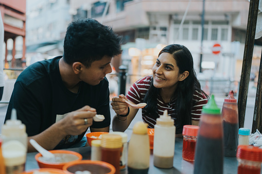 A medium shot of a young foreign tourist couple enjoying Hong Kong local food. They are looking at each other happily as they are about to try the rice in the spoons. Multiple small orange bowls with different delicacies and sauces are scattered around the table