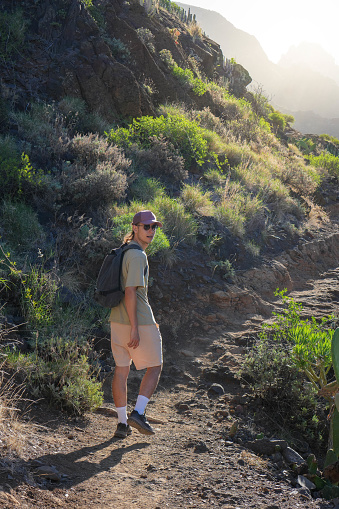 vertical frame of solitary hiker, viewed from behind, ascending a rugged mountain trail at sunrise. With high socks, shorts, a t-shirt, and a backpack. The surrounding terrain is wild and rocky, with shrubs lining the trail. The early morning sun filters through the mountains, casting a warm, atmospheric glow on the scene.