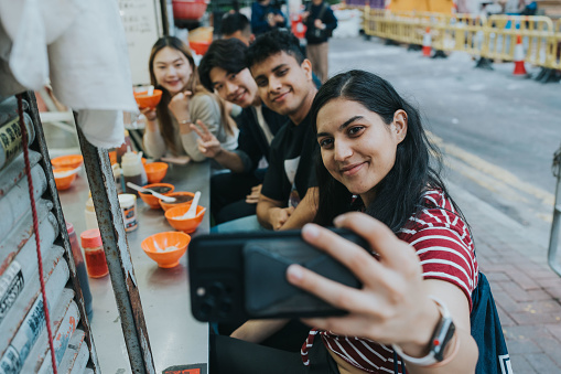A medium shot of a young Indian woman holding out her hand with her phone to take a selfie photo of her and her international friends. Her foreign friends are smiling and posing for the picture. They are eating local street food served in small orange bowls. This is a cozy experience to remember their moments together