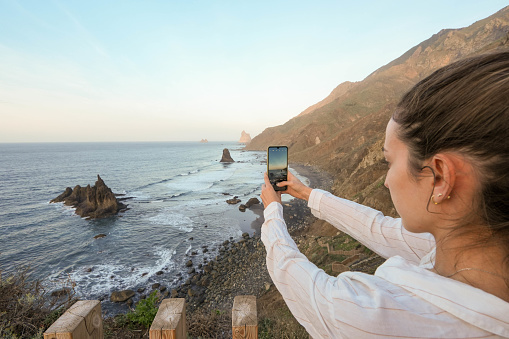 An adult in a white shirt captures the scenic ocean view, featuring rocky outcrops and waves, during the golden hour using a smartphone
