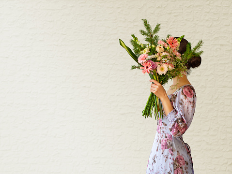 Woman in a purple dress holding a bouquet of flowers in front of her face. Isolated white wall background. Graduation, celebration, surprise event concept. Copy space.