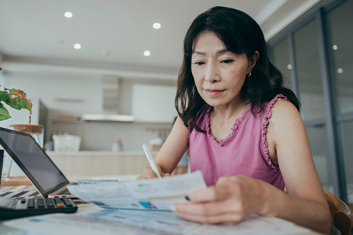 A woman is at home, holding a collection of paper bills and financial plans. She appears to be focused on managing her personal finances, possibly sorting through bills, budgeting, or organizing her financial matters.