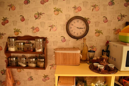 part of the kitchen wall with antique decorative wallpaper a wooden shelf with spices and a wall clock a shelf with a bread box, a bottle and a glass and small coffee cups