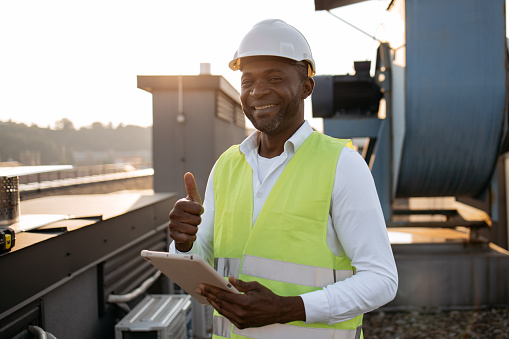 Serious factory worker standing and holding modern tablet in hands while servicing devices at factory. African american man wearing uniform inspecting area while performing work in local roof.